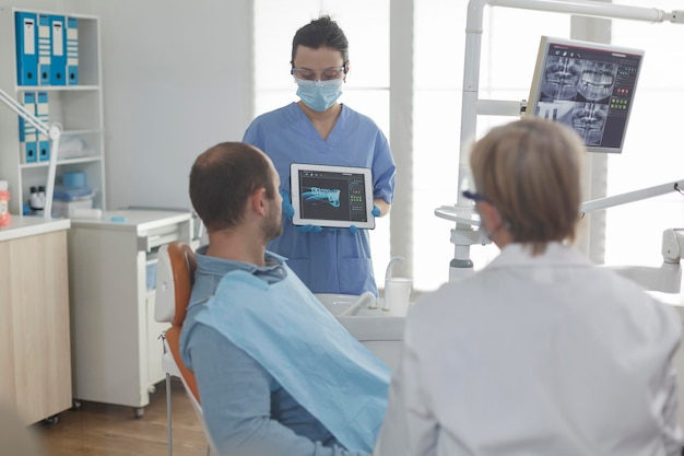 Assistente dentista che indossa una maschera facciale che mostra la radiografia dei denti utilizzando un tablet al paziente che discute della procedura medica durante l'esame stomatologico nello studio dentistico. Uomo malato con infezione orale