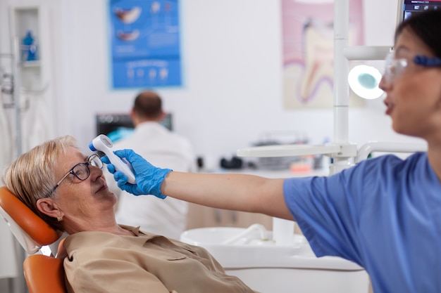 Assistente dentista che tiene l'indicatore digitale della temperatura corporea davanti al paziente seduto sulla sedia