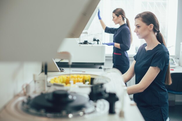 Assistente di laboratorio allegro che sorride mentre è seduto e il suo posto di lavoro. Collega concentrato che apre un analizzatore biochimico sullo sfondo