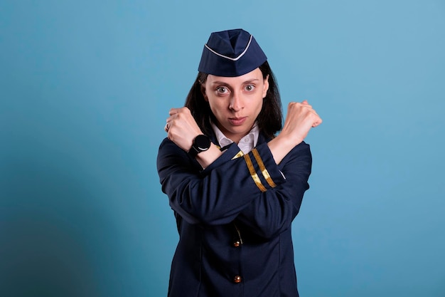 Assistente di volo che mostra un gesto di arresto con le braccia incrociate, i pugni chiusi. Hostess in uniforme che guarda la fotocamera con seria espressione facciale, concetto di divieto, simbolo di difesa