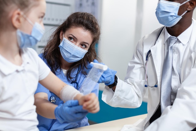 Assistente medico che aiuta il medico pediatra afroamericano a fasciare il braccio rotto del paziente del bambino piccolo durante la fisioterapia clinica nell'ufficio dell'ospedale. Squadra con mascherina contro il coronavirus