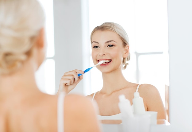 assistenza sanitaria, igiene dentale, persone e concetto di bellezza - giovane donna sorridente con spazzolino da denti che pulisce i denti e cerca di specchiarsi nel bagno di casa