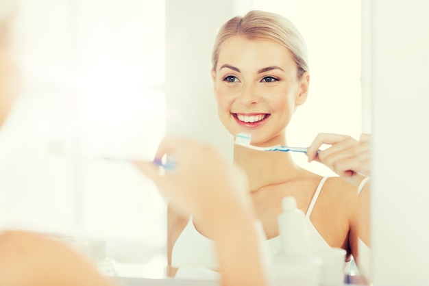 assistenza sanitaria, igiene dentale, persone e concetto di bellezza - giovane donna sorridente con spazzolino da denti che pulisce i denti e cerca di specchiarsi nel bagno di casa