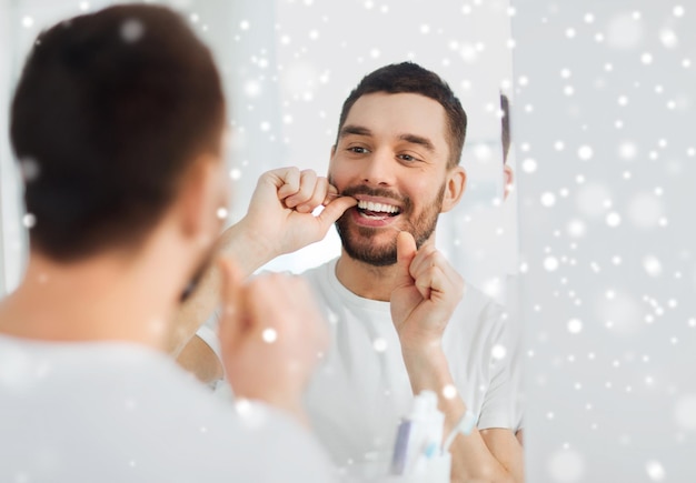 assistenza sanitaria, igiene dentale, persone e concetto di bellezza - giovane sorridente con il filo interdentale che pulisce i denti e cerca di specchiarsi nel bagno di casa sulla neve