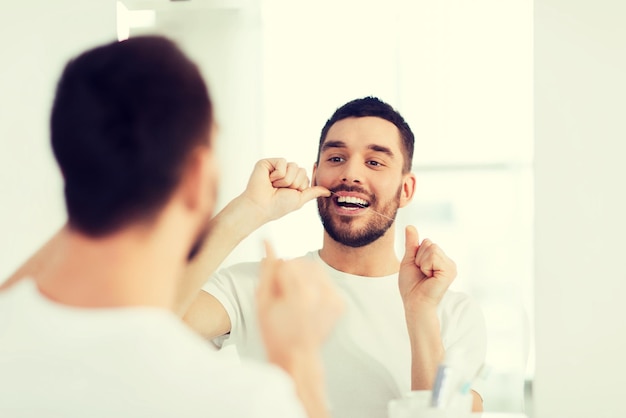 assistenza sanitaria, igiene dentale, persone e concetto di bellezza - giovane sorridente con il filo interdentale che pulisce i denti e cerca di specchiarsi nel bagno di casa