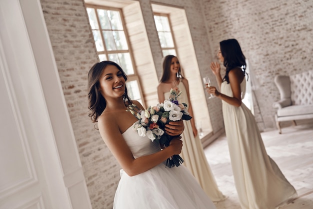 È assolutamente pronta. Attraente giovane donna con in mano un bouquet da sposa e sorridente mentre le sue amiche bevono champagne sullo sfondo