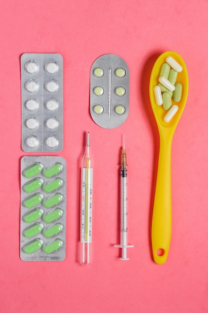 Assortimento di pillole e capsule in cucchiaio giallo, siringa, termometro sul rosa.