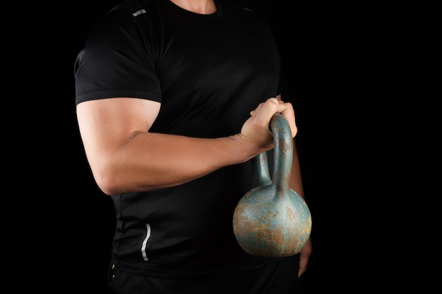 Atleta adulto forte in abiti neri con un kettlebell di ferro sulle braccia tese