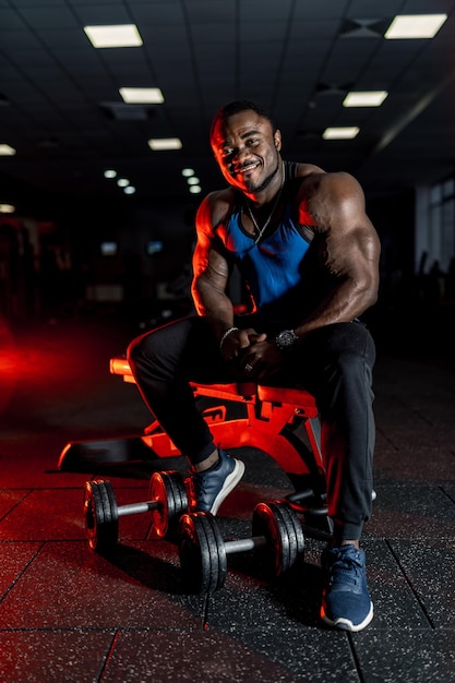 Atleta africano molto muscoloso si siede sulla panchina in palestra moden. Lo sport come stile di vita. Luce oscura. avvicinamento.