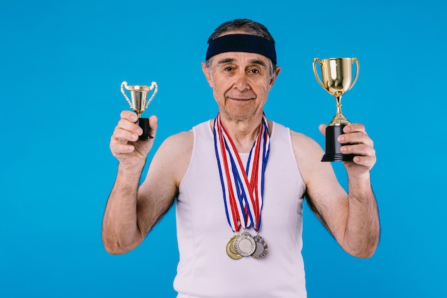 Atleta anziano con segni di sole sulle braccia, con tre medaglie al collo e due trofei in mano, su sfondo blu