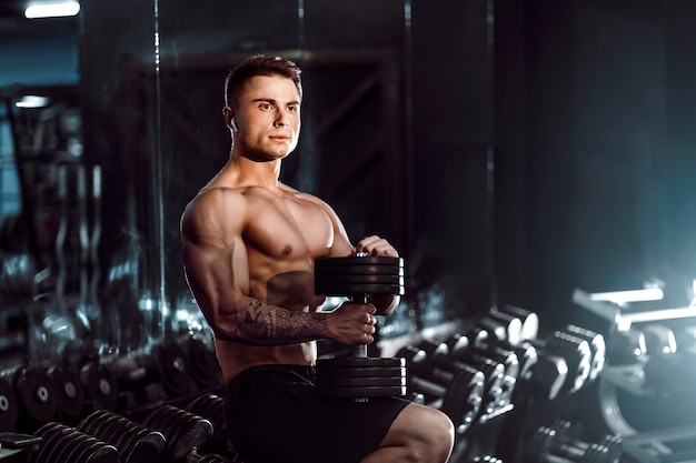 Atleta bello del giovane adulto che risolve in palestra, sedendosi su una panchina e tenendo la testa di legno con le braccia alzate. al coperto, guardando peso