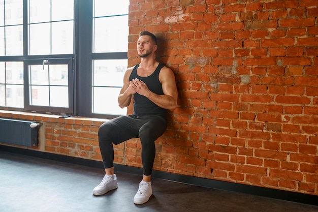 Atleta che fa esercizio di formazione contro la parete della palestra. Sportivo che esercita squat appoggiandosi indietro sul muro di mattoni. Concetto di sport, bodybuilding, fitness e persone