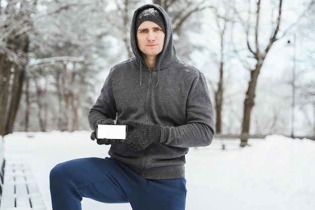 Atleta del giovane che mostra smartphone con uno schermo vuoto durante l'allenamento invernale nel parco cittadino innevato