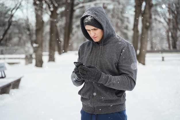 Atleta del giovane che utilizza smartphone durante l'allenamento invernale nel parco cittadino innevato