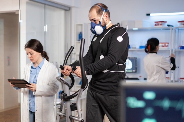 Atleta di prestazioni in esecuzione su cross trainer con elettrodi attaccati sul corpo e maschera nel laboratorio di test, medico scienziato che monitora la frequenza cardiaca guardando il tablet pc.