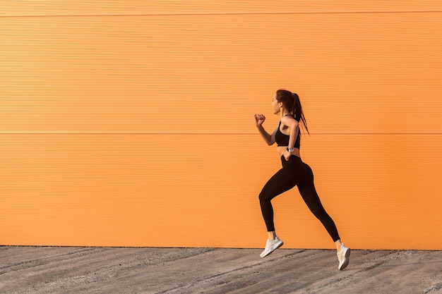 Atleta donna motivata in forma sicura in abbigliamento sportivo attillato, pantaloni neri e top, iniziando a correre, fare jogging all'aperto contro il muro arancione, area pubblicitaria. Assistenza sanitaria e dimagrimento, attività sportiva