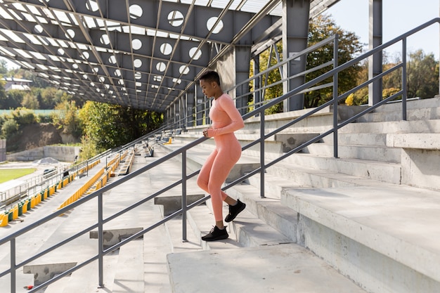 Atleta femminile che fa fitness in una tuta rosa vicino a uno stadio sportivo, afroamericana che corre su per le scale al mattino