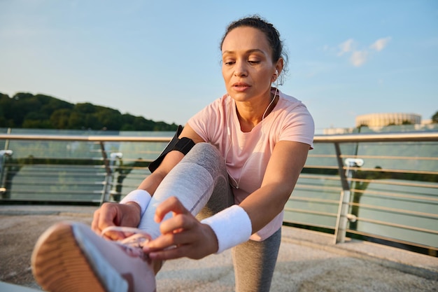 Atleta femminile con auricolari, corridore che lega i lacci delle scarpe da ginnastica mentre si gode la corsa mattutina su un tapis roulant del ponte della città. Corri, jogging, stile di vita attivo sano, allenamento cardio e concetto di fitness all'aperto