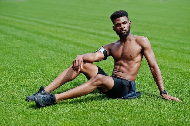 Atleta maschio afroamericano sport sexy torso nudo uomo con custodia per braccio sportivo in esecuzione per telefono cellulare posato sull'erba verde dello stadio di calcio