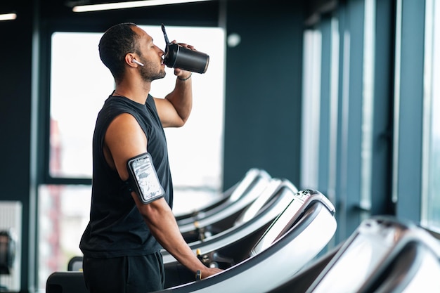 Atleta maschio nero bello che beve acqua durante l'allenamento al tapis roulant in palestra