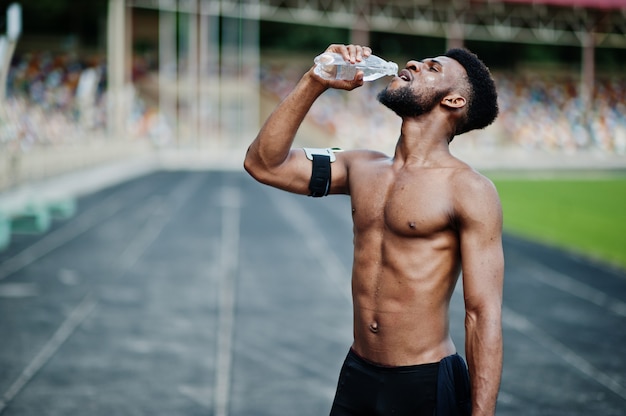 Atleta maschio senza camicia con custodia da braccio sportiva per telefono cellulare, posato allo stadio e acqua potabile dalla bottiglia