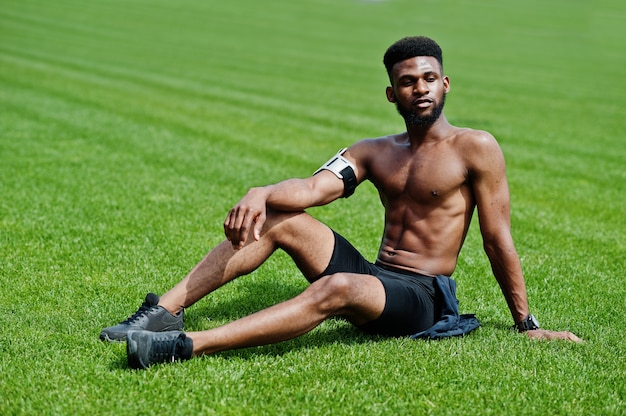 Atleta maschio senza camicia con la pratica del braccio sportivo per il telefono cellulare, posato ad erba verde dello stadio di calcio