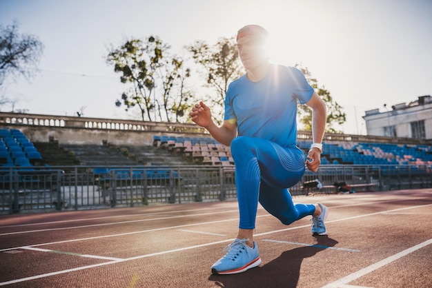 Atleta muscolare giovane maschio che allunga la gamba su una pista da corsa nello stadio preparandosi per correre e fare jogging Uomo caucasico che esercita attività all'aperto indossando abbigliamento sportivo blu Sport e persone
