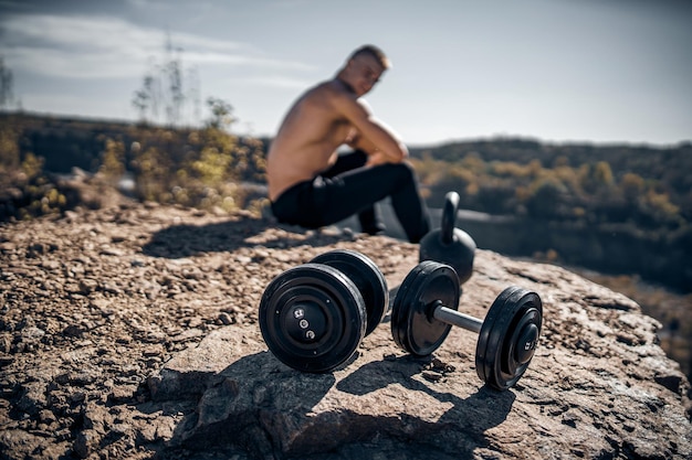 Atleta seduto su una roccia Paesaggio naturale Dumbbels on front vew Sfondo sfocato