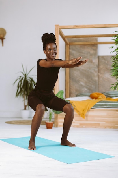 Atletica donna afroamericana con sorriso accovacciato con le braccia estese in avanti allenamento sul tappetino ginnico in studio decorato Pompaggio muscolare esercizi sportivi fitness mantenendo il corpo in forma