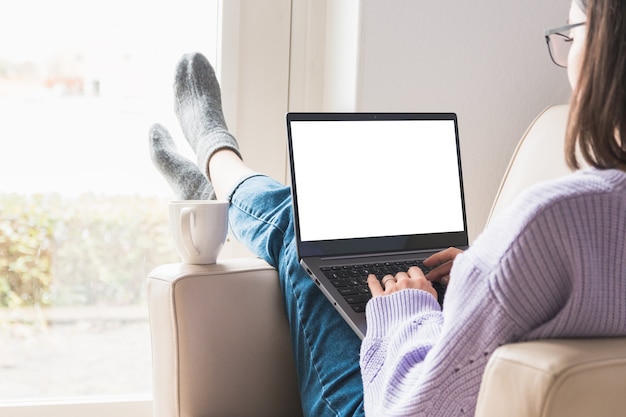 Atmosfera accogliente lavorando e studiando a casa. vista laterale di una rilassata attraente giovane donna professionale o studente con gli occhiali, lavorando su un computer in poltrona. schermo bianco.