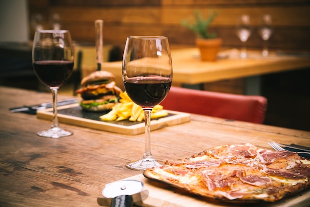 Atmosfera tradizionale del ristorante con pizza al prosciutto e hamburger con patatine sullo sfondo su tavola in legno rustico e bicchiere di vino rosso