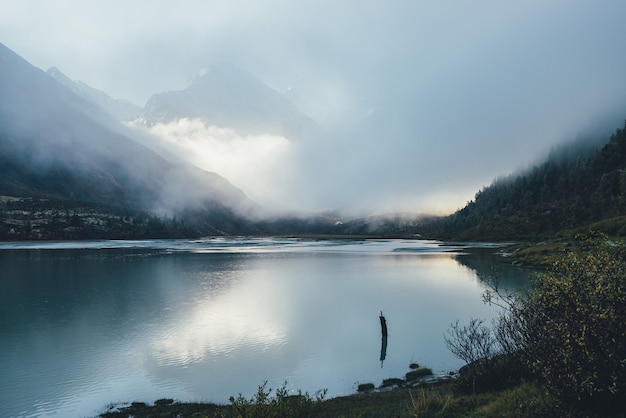 Atmosferico paesaggio alpino con lago di montagna e alta montagna innevata in dense nuvole basse. Bellissimo scenario di scarsa illuminazione con picco appuntito in una fitta nebbia e riflesso del sole dorato nel lago.