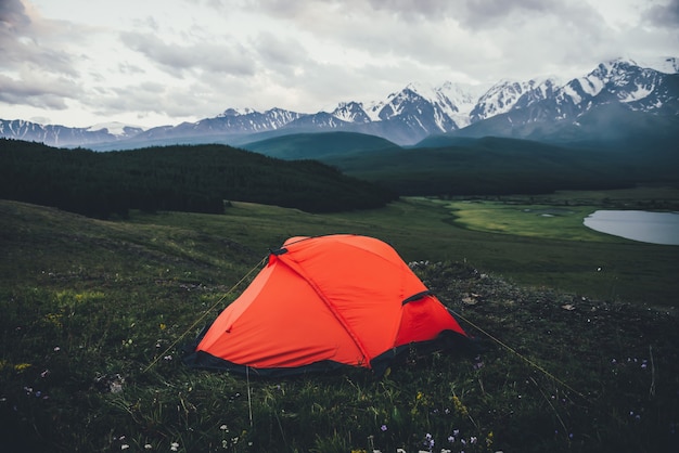 Atmosferico paesaggio alpino con tenda arancione sullo sfondo del lago e grandi montagne innevate con tempo nuvoloso. Fantastico scenario verde con tenda arancione con vista sulle grandi montagne sotto il cielo nuvoloso.