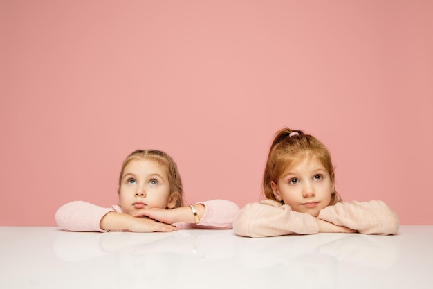 Attento. Bambini felici, ragazze isolate su sfondo rosa corallo per studio. Sembra felice, allegro. Copyspace per l'annuncio. Infanzia, educazione, emozioni, concetto di espressione facciale. Seduto a tavola, carino.