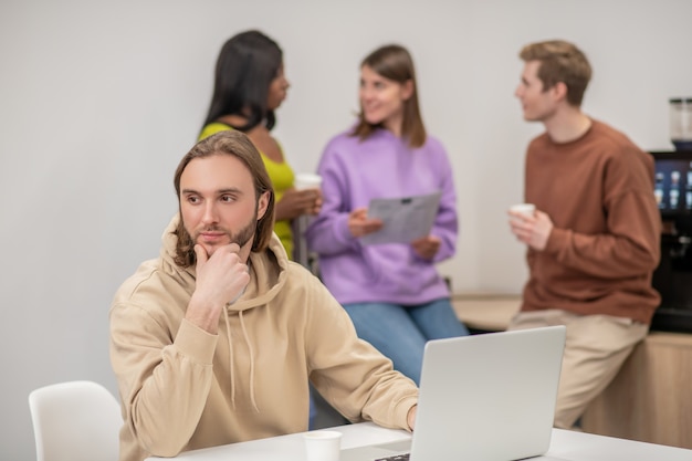 Attivismo, movimento. Pensando ragazzo ottimista seduto vicino a laptop e colleghi di giovani organizzatori in piedi dietro parlando di importante