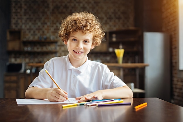 Attività creativa. Ritratto di un giovane diligente seduto a un tavolo di legno e guarda nell'obiettivo con un sorriso allegro sul viso mentre lavora a un nuovo capolavoro con matite colorate.