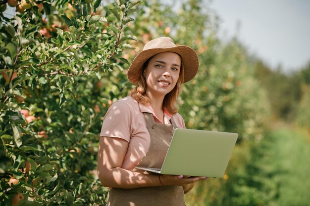 Attraente agronomo o agricoltore femminile con laptop in piedi nel frutteto di mele e il controllo della frutta prende appunti Concetto di agricoltura e giardinaggio Alimentazione sana