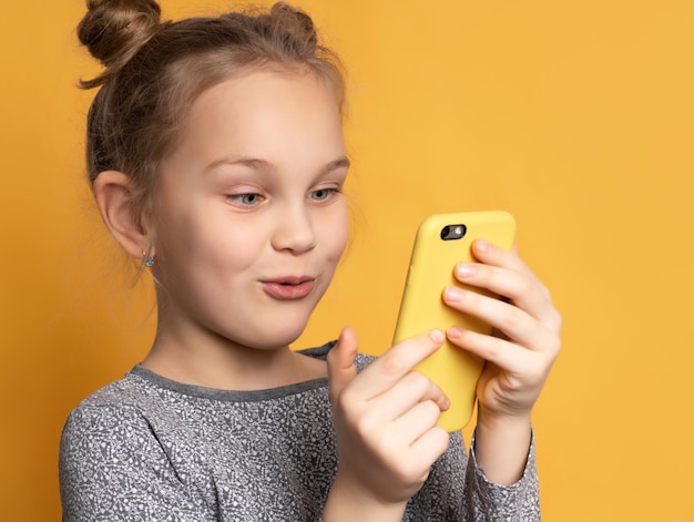 Attraente bambina bionda che guarda lo schermo del suo telefono cellulare con un viso gioioso, su sfondo giallo per lo spazio della copia