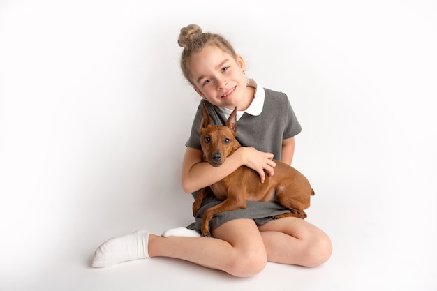 Attraente bambina in un abito da scuola rigoroso con un pinscher marrone nano su sfondo bianco ammira un'espressione felice sul viso. Foto di alta qualità