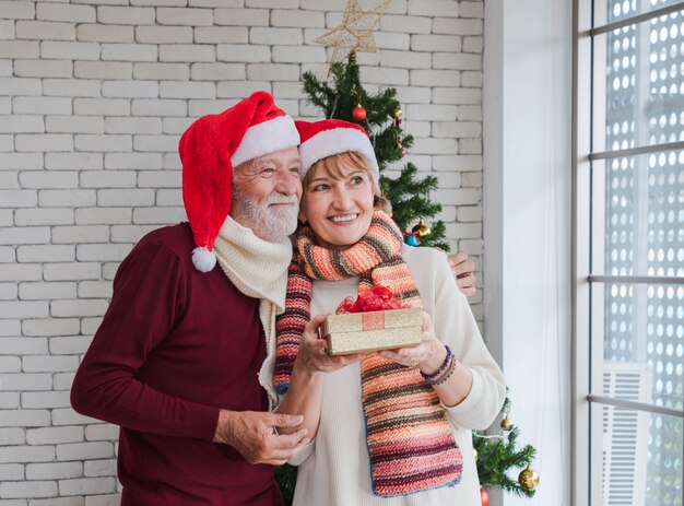 Attraente coppia di anziani caucasici che si abbracciano o si abbracciano con amore e guardano avanti con umore positivo e speranza per una nuova vita durante la celebrazione del Natale e del nuovo anno insieme al presente in mano.