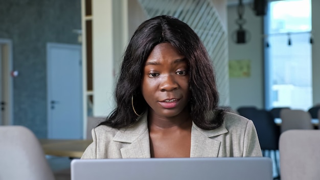 Attraente donna afro-americana positiva parla in videochat tramite laptop contemporaneo seduto al tavolo in un accogliente ristorante vista ravvicinata