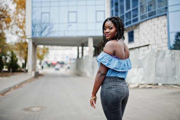 Attraente donna afroamericana con dreadlocks in jeans indossano poste contro un moderno edificio a più piani