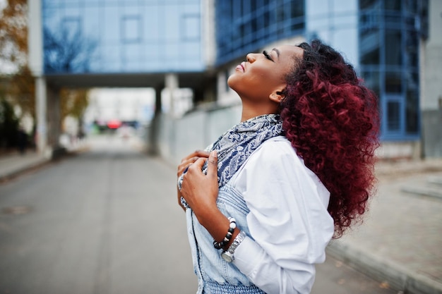 Attraente donna afroamericana riccia in abito jeans poste contro un moderno edificio a più piani