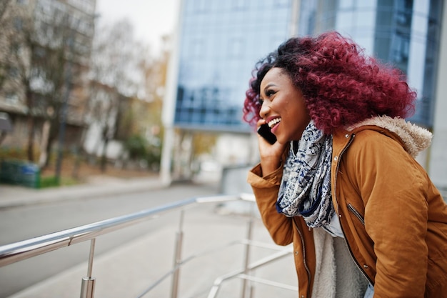 Attraente donna afroamericana riccia in cappotto marrone posata vicino alle ringhiere contro un moderno edificio a più piani che parla sul telefono cellulare