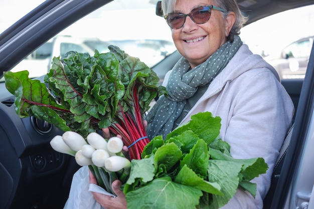 Attraente donna anziana entra in macchina dopo aver acquistato verdure fresche al mercato degli agricoltori. Ha in mano lattuga verde e cipollotti. Concetto di stile di vita sano e vegetariano