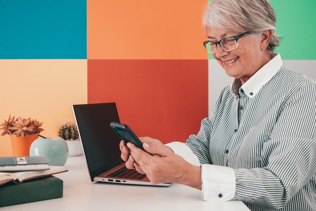 Attraente donna anziana in camicia e occhiali seduto alla scrivania dell'ufficio utilizzando lo smartphone