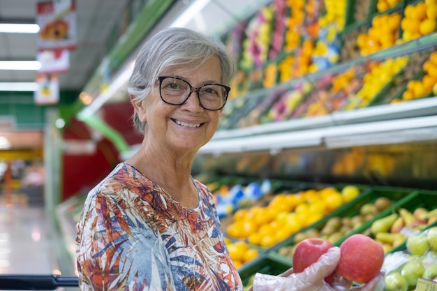 Attraente donna anziana in un supermercato che sceglie frutta fresca con due mele rosse nelle mani. Frutta mista colorata su sfondo
