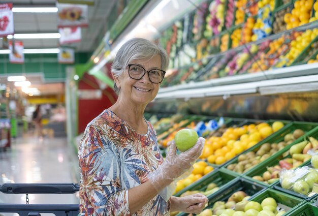 Attraente donna anziana in un supermercato che sceglie frutta fresca, con in mano una mela verde. Frutta mista colorata su sfondo