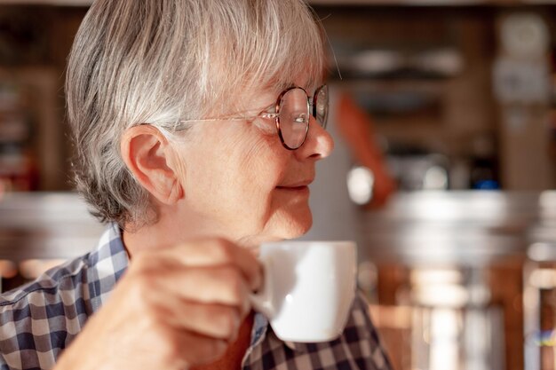 Attraente donna anziana seduta al coperto al tavolino di un bar che si gode una tazza di caffè espresso signora anziana caucasica con gli occhiali rilassati ha una pausa tenendo una tazza di caffè