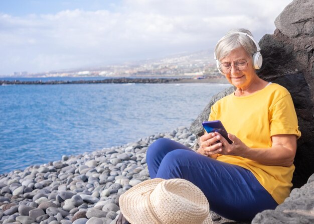 Attraente donna anziana seduta su una spiaggia di ciottoli in mare utilizzando il telefono cellulare Donna matura sorride tenendo il cellulare ascoltando musica con le cuffie Concetto di libertà di vacanza e felice pensionamento